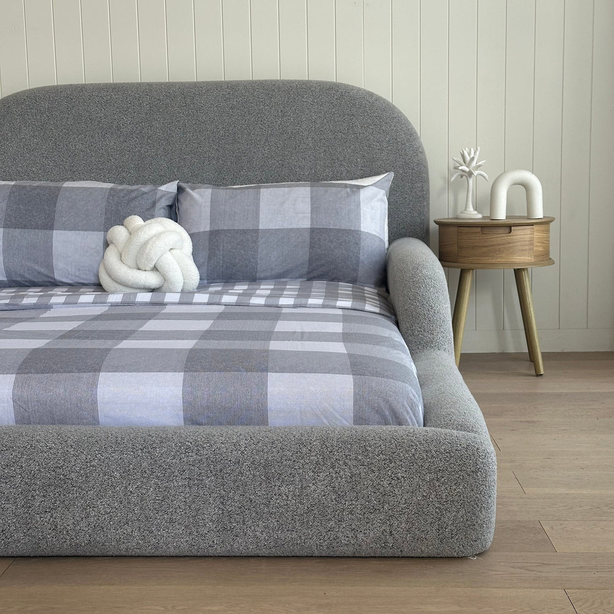 Gray textured headboard with plaid blanket and white decorative item against a wooden wall.