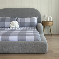 Gray textured headboard with plaid blanket and white decorative item against a wooden wall.