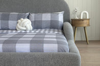 Gray textured headboard with plaid blanket and white decorative item against a wooden wall.