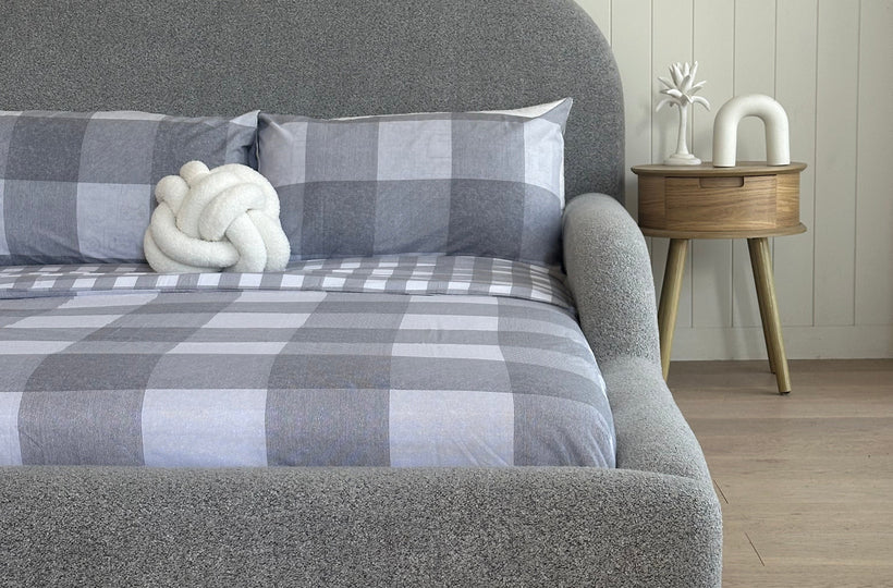 Gray textured headboard with plaid blanket and white decorative item against a wooden wall.